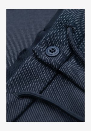 Gros plan d'un pantalon en tissu texturé bleu foncé avec un bouton portant la mention "OMBRE" et un cordon apparent à la taille élastique.