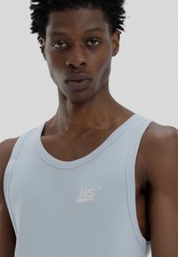 Camisola de alças larga, em azul claro, com textura canelada. Apresenta um pequeno logótipo em branco bordado no peito. Design ajustado e textura suave.