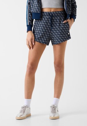 Femme portant un short bleu marine à motif, un crop top gris, une veste bleu marine, des chaussettes blanches et des baskets argentées, debout avec une main dans la poche.