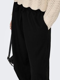 Άτομο που φορά χαλαρό μαύρο παντελόνι με κορδόνι στη μέση και μπεζ πλεκτό πουλόβερ με υφή, κρατώντας λουρί μαύρης τσάντας με πόρπη σε σχήμα δαχτυλιδιού.