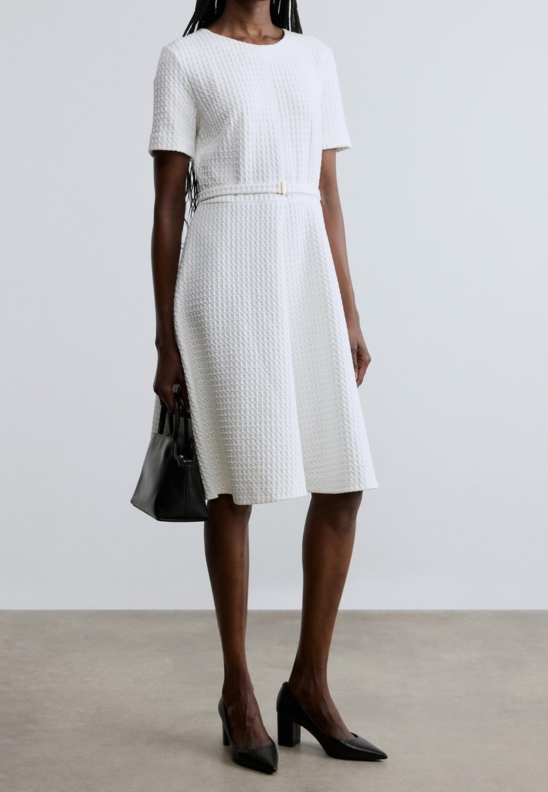 Femme portant une robe blanche texturée midi avec des manches courtes et une fine ceinture, tenant un sac à main noir et portant des talons hauts noirs.