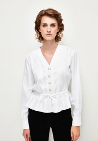 Blusa bianca con scollatura a V, chiusura frontale con bottoni e vita con coulisse. Presenta dettagli a pin tuck e maniche lunghe. Tessuto di cotone liscio.