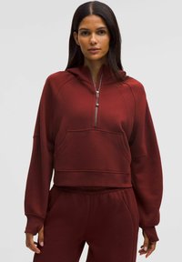 Hoodie cropped di un rosso intenso con tasca frontale, colletto con zip e tessuto testurizzato. Presenta maniche ampie e un design minimalista.