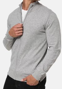 Šedý svetr na zip s dlouhými rukávy, žebrovaným límcem a manžetami a hladkou texturou. Má přiléhavý střih, nošený přes bílou košili.