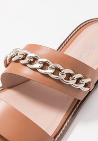 Bruine leren slipper met een brede band en een decoratief ontwerp van een zilveren metalen ketting. Glad oppervlak met een lichtroze binnenzool.