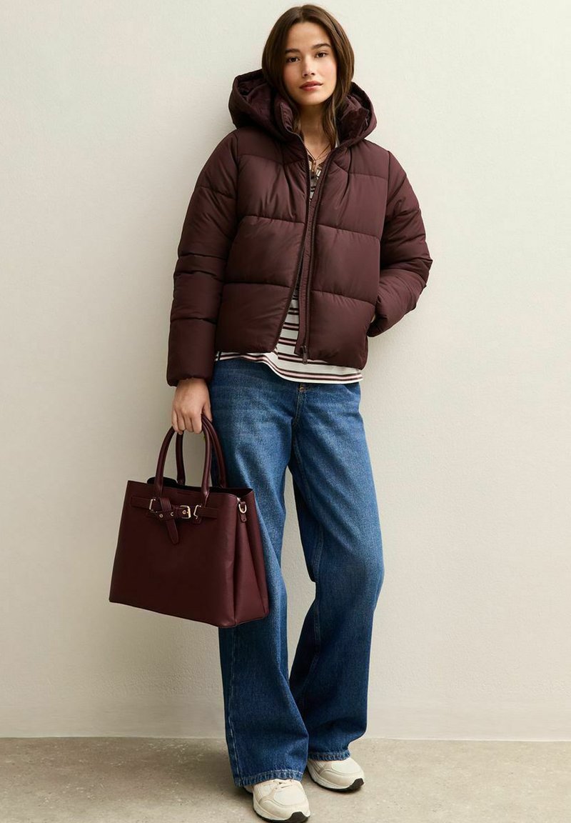 Giacca puffer bordeaux con cappuccio, chiusura con zip e design trapuntato. Abbinata a una maglietta a righe e jeans blu a gamba larga. Tiene una borsa bordeaux.