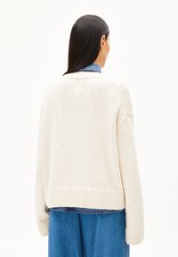 Chunky gestrickter cremefarbener Pullover mit Rippensaum und -bündchen, in lockerer Passform. Darüber getragen wird ein blauer Denim-Hemd.