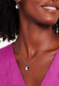 Yin-yang hanger ketting aan een zilveren ketting, vergezeld van bijpassende yin-yang oorbellen. De achtergrond heeft een gestructureerde paarse stof.