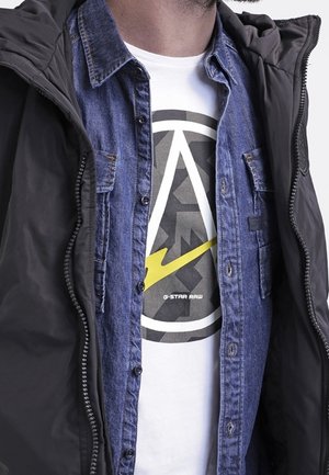 Blue denim shirt layered under a black zippered jacket, featuring a graphic T-shirt with a circular logo in grey and yellow lightning bolt design.