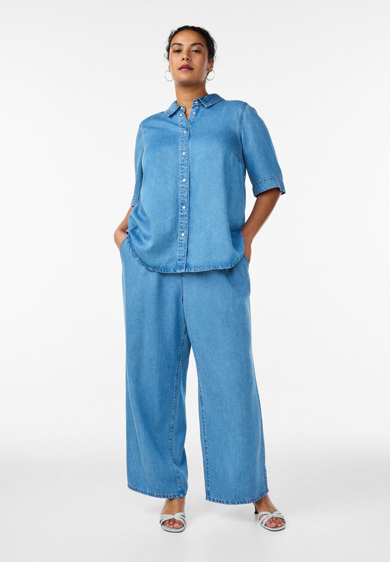 Kvinde stående med hænder i lommerne iført lyseblå denimskjorte med halvlange ærmer, vide jeans og hvide åbne sandaler på hvid baggrund.