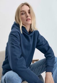 Felpa blu navy con una texture morbida, dotata di cappuccio con lacci, polsini a coste e vestibilità rilassata, abbinata a jeans blu chiaro.