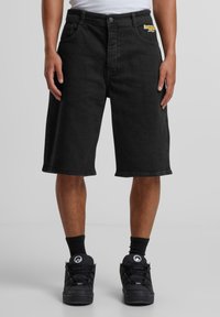 Pantalones cortos de mezclilla negra con un ajuste relajado, bolsillos frontales y un sutil logo bordado. Combinados con zapatillas deportivas negras y calcetines.