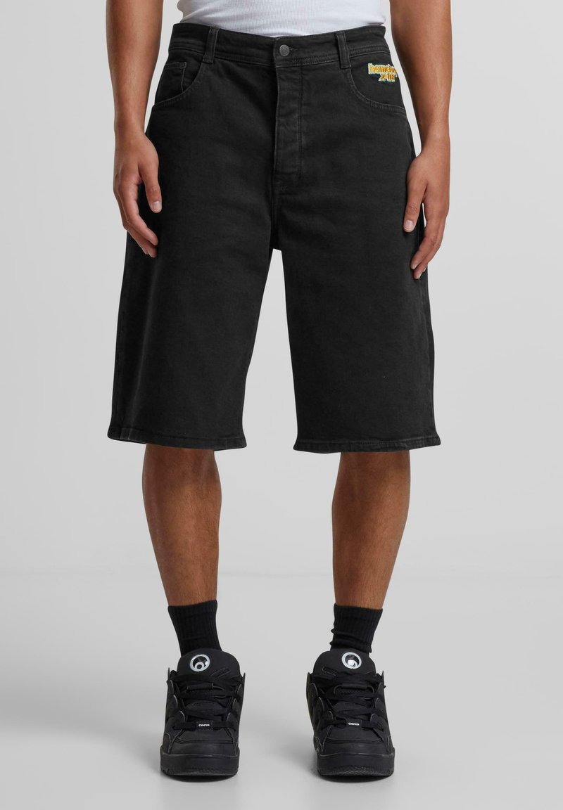 Pantalones cortos de mezclilla negra con un ajuste relajado, bolsillos frontales y un sutil logo bordado. Combinados con zapatillas deportivas negras y calcetines.