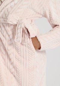 Accappatoio rosa chiaro in materiale peluche, con un motivo a treccia testurizzata, vita con cintura e tasche su entrambi i lati.
