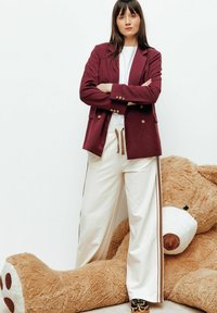 Blazer double boutonnage bordeaux sur un t-shirt blanc, associé à un pantalon de survêtement ivoire avec des rayures marron sur les côtés, le tout posé devant un énorme ours en peluche.