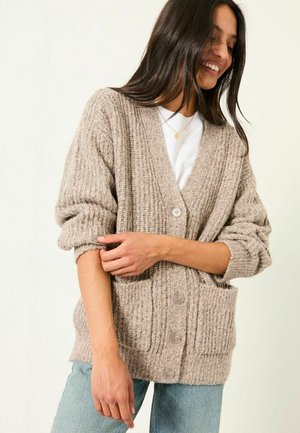 Beige ribbed cardigan with a V-neck, large buttons, and two front pockets. Knit texture with rolled sleeves, worn over a white shirt.