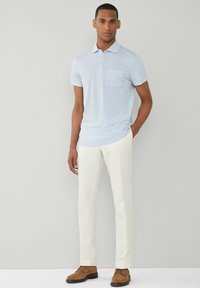 Svetlomodrá polo tričko s vreckom na hrudi, skombinované s bielymi nohavicami a hnedými semišovými topánkami. Jednoduchý, moderný dizajn s jemnou textúrou.