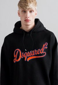Junger blonder Mann in einem schwarzen Hoodie mit dem orangefarbenen und blauen "Dsquared²" Logo auf der Vorderseite vor einem schlichten, hellen Hintergrund.