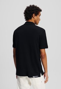 Μαύρο πόλο μπλουζάκι με κοντά μανίκια, γιακά και πλαϊνές σχισμές, κατασκευασμένο από λείο ύφασμα. Διαθέτει διακριτικό λογότυπο στο τελείωμα.