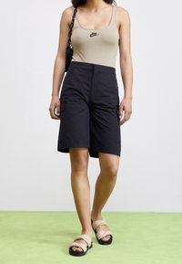 Beige linne med svart Nike-logotyp; svarta cargoshorts med fickor; bruna sandaler med knutna remmar; modell som står framför en grön bakgrund.