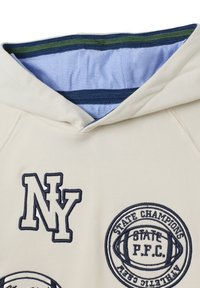 Haut de sweatshirt beige clair avec le logo brodé "NY" bleu marine et un emblème sportif. L'intérieur présente une doublure bleue avec des rayures vertes et bleu marine.