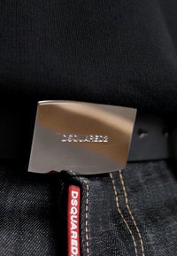 Detailní záběr na černý kožený opasek se stříbrnou sponou s vyrytým nápisem "DSQUARED2" a denimové džíny s červenou etiketou "DSQUARED" na kapse.