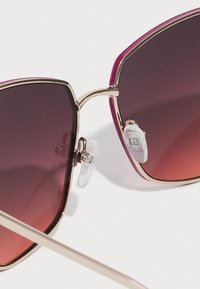 Sunglasses with a geometric, oversized shape. Gradient purple lenses, gold metal frame, and clear nose pads. Thin, light-colored temples.