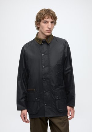 Junger Mann mit lockigem Haar, der eine schwarze Jacke mit braunem Cordkragen und mehreren Fronttaschen trägt, steht vor einem schlichten Hintergrund.