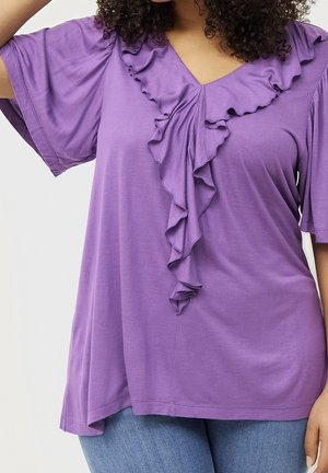 Woman wearing a loose purple blouse with ruffled V-neck detail and short sleeves, paired with blue jeans, standing against a plain background.