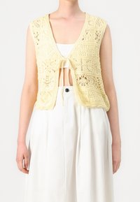 Neulotut lyhyet liivi vaaleankeltaisena, avoimella etuosalla, monimutkaisilla pitsikuvioilla ja solmittavalla sulkemisella, kerroksittain valkoisen topin päällä.