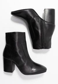 Par de botines de cuero negro con tacones cuadrados y cremalleras laterales, exhibidos sobre un fondo blanco.