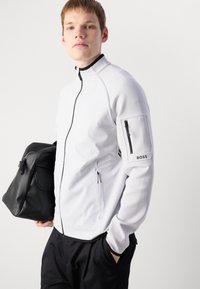 Jachetă albă cu fermoar, cu accente negre, țesătură texturată, buzunare laterale și un buzunar pe braț cu un logo. Modelul ține o geantă neagră.