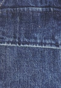 Denimstof i en dyb blå farve med en glat tekstur. Billedet viser en tæt på vew af stofvævningen og syningsdetaljerne.