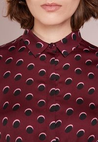 Blusa bordeaux con colletto caratterizzata da un motivo di forme a mezzaluna alternate in nero e rosa. Tessuto morbido con chiusura a bottone al colletto.