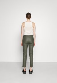 Olijfgroene leren-look broek met slim-fit ontwerp, iets uitlopend aan de onderkant. Draagt een crème tanktop en zwarte plateau schoenen.