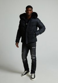 Svart dunjakke med pelsforet hette, med kviltet tekstur, glidelås og sidelommer, paret med slitte mørke jeans og joggesko.