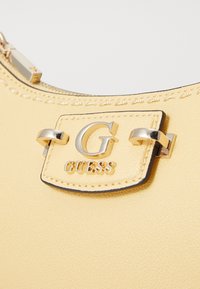 Sac à main synthétique jaune avec une surface texturée, quincaillerie en ton doré et une plaque de marque avec la lettre "G" et "GUESS" en relief.