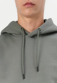 Hooded sweatshirt in muted green, made of soft fabric. Features drawstrings with black tips and a textured surface.