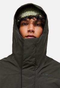 Jachetă impermeabilă de un verde închis, cu guler înalt și glugă mare. Căciulă tricotată texturată în tonuri de verde sub glugă. Brand minimal prezent.