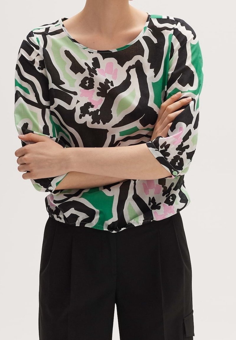 Personne portant un chemisier floral abstrait coloré avec des motifs verts, noirs et roses et un pantalon noir taille haute, les bras croisés.