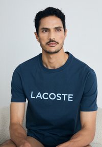 Tamno plava pamučna majica s kratkim rukavima koja sadrži "LACOSTE" logo u svijetloplavoj boji preko prsnog dijela. Mekan na dodir i klasičnog kroja.