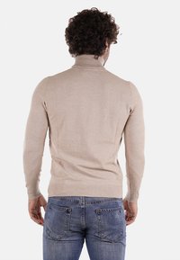 Maglione a collo alto color beige chiaro in tessuto morbido, design aderente, con polsini e orlo a coste. Indossato con jeans blu, visto da dietro.