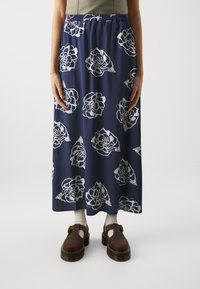 Mørkeblå maxiskjørt med hvit blomstermønster, elastisk midjebånd, lett stoff og flytende design. Kombinert med brune sko og sokker.