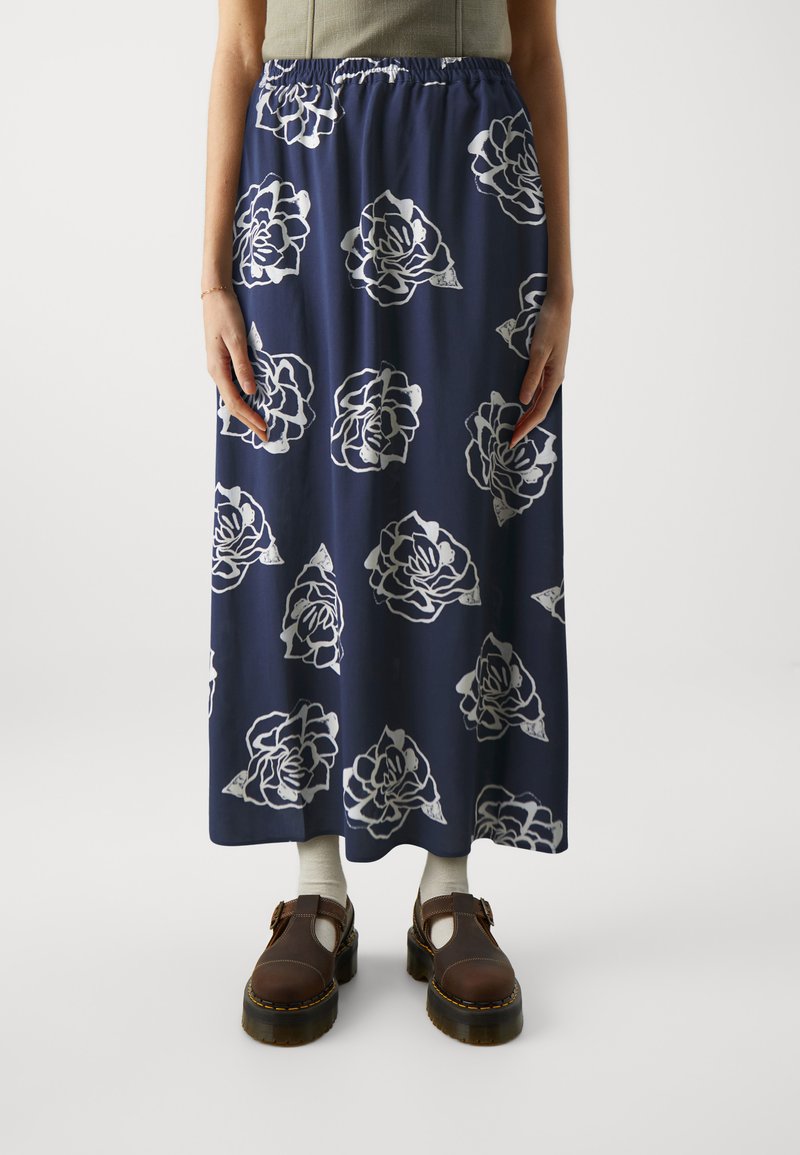 Mørkeblå maxiskjørt med hvit blomstermønster, elastisk midjebånd, lett stoff og flytende design. Kombinert med brune sko og sokker.