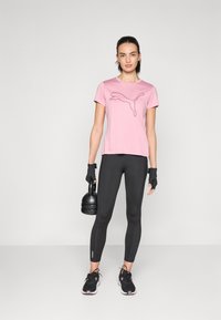 Lys rosa treningst-skjorte med svart Puma-logo, kombinert med svarte tights og svarte joggesko. Personen holder en svart kettlebell og har på seg hansker.