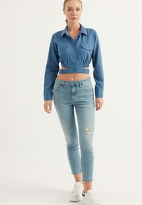 Camicia blu a maniche lunghe con due tasche sul petto, abbinata a jeans skinny chiari strappati e sneaker bianche.