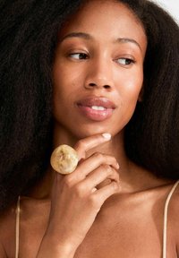 Kollane vaigust sõrmus ümarate, ebaühtlaste servadega, millel on poleeritud ja sile pind ning hele marmormuster. Käsi puhkab õrnalt lõual.