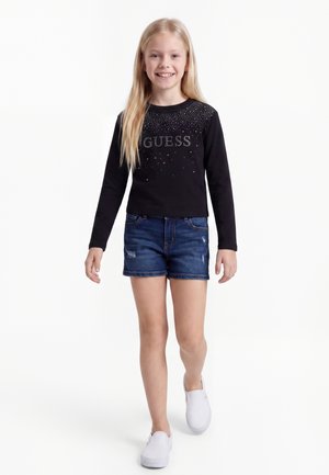 Ung pige med langt blondt hår har en sort langærmet skjorte med teksten "GUESS", denimshorts og hvide slip-on sko på og går fremad smilende.