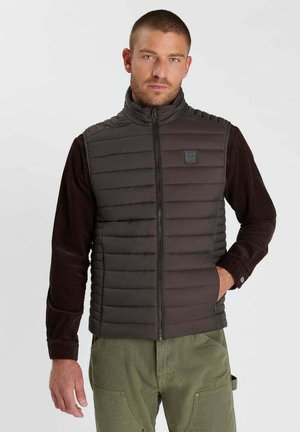 Hombre que lleva un chaleco acolchado sin mangas de color marrón oscuro sobre una camisa de manga larga oscura y pantalones verde oliva, de pie con una mano en el bolsillo.