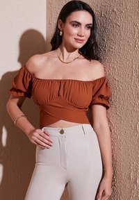 Rustrød off-shoulder top med puffærmer og sammensyninger, parret med højremsbeige bukser med en frontlukning med knap.